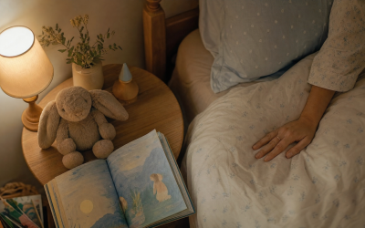 Mesilla infantil con lamparita encendida, peluche y cuento, con la mano de una madre sobre la cama - blog de Clínica Lazos